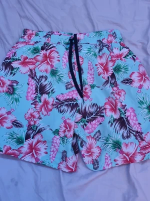 Blommiga turkosa badbyxor med snörning - Säljer ett par badbyxor med turkos botten och färgglatt blommönster i rosa, grönt och brunt. De har svart snörning i midjan och en bakficka med knapp. Perfekta för stranden eller poolen!