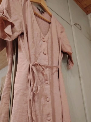 Ljusrosa klänning med knytärm🩷  - Supersöt ljusrosa klänning med v-ringning, knappar framtill och ett matchande bälte i midjan🩷 Klänningen har korta ärmar med knyt och är gjord i ett lätt linnematerial som passar perfekt till varmare dagar🌸☀️ Passar storlek S. 