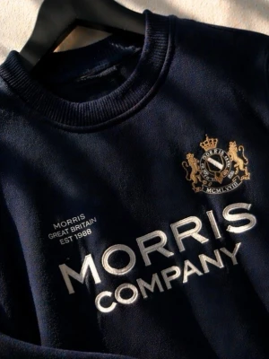 Mörkblå sweatshirt från Morris Company - Mörkblå sweatshirt från Morris Company med broderad logga och text på bröstet. Tröjan har rund halsringning, ribbade muddar och detaljer i guld och vitt. Tillverkad i mjukt material som känns skönt mot huden. Perfekt för en stilren och avslappnad look.