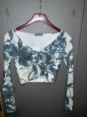 Blommig croppad topp från Guess - Supersnygg croppad topp från Guess med blått blommönster på vit botten. Toppen har vringning, långa ärmar och en liten metalllogga vid nederkanten. Materialet är ribbad bomull som känns mjuk och följsam. Perfekt för dig som vill sticka ut lite extra!