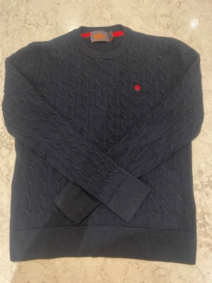 Mörkblå kabelstickad tröja Morris - Snygg mörkblå kabelstickad tröja från Morris i 100% merinoull. Tröjan har rund halsringning, ribbade muddar och en liten röd broderad logga på bröstet. Perfekt för dig som gillar klassisk och stilren design. Den har dock krympt lite