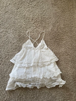Vit volang klänning med spets - Supersöt vit linnetopp med tunna axelband och flera lager volanger. Toppen har v-ringning och fina spetsdetaljer längs kanterna som ger en romantisk och luftig känsla. Perfekt för sommarens alla dagar.