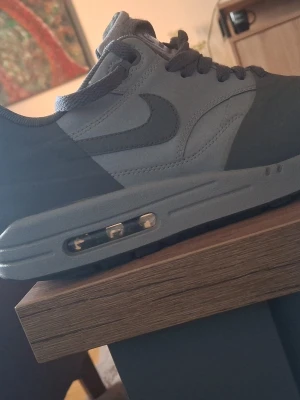 Grå Nike Air Max 1 sneakers - Säljer ett par grå Nike Air Max 1 sneakers med klassisk låg profil och synlig Air-dämpning i sulan. Skorna har en kombination av mocka och textilmaterial, ikonisk Swoosh på sidan och matchande grå skosnören. Perfekt för dig som gillar stilrena och sportiga sneakers.