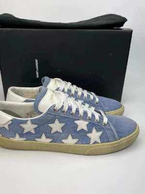 Saint Laurent blå stjärnsneakers - Snygga sneakers från Saint Laurent i ljusblå denim med vita stjärnor på sidorna. Skorna har vita snören, vit häl och beige sula. Märkeslogga på plösen och klassisk låg modell som ger en cool och unik look.