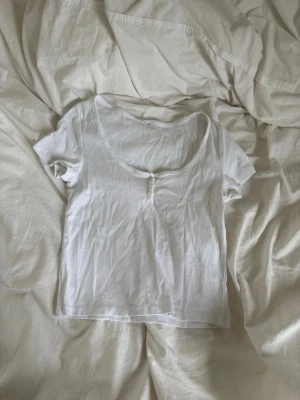 Vit topp med knappar och rund hals - En enkel vit topp med korta ärmar och rundad halsringning. Framtill finns små dekorativa knappar som ger en snygg detalj. T-shirten är gjord i mjukt bomullsmaterial och har möjlighet att välja urringningen beroende på hur många knappar du knäpper 