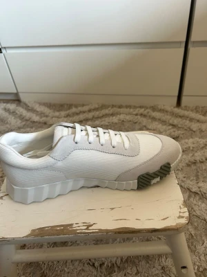 Vita H sneakers med grå detaljer - Snygga vita sneakers med grå detaljer och ribbad sula. Ovandelen är i textil och mocka, med vita skosnören och en unik grå detalj på sulan. Perfekt för dig som gillar stilrena och moderna skor.