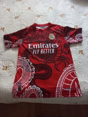 Röd Real Madrid matchtröja drakmotiv - Säljer en röd Real Madrid matchtröja från Adidas med ett stort svart och vitt drakmotiv över hela tröjan. Tröjan har korta ärmar, rund hals och klubbmärke på bröstet. Emirates Fly Better-logga tryckt framtill. Perfekt för fotbollsfans!