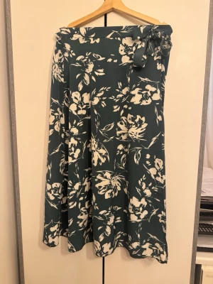 Grön blommig omlottkjol med knyt - Snygg långkjol i mörkgrönt med vitt blommönster. Kjolen har omlottmodell och knyts i midjan för en justerbar passform. Perfekt för dig som gillar en flowy och avslappnad stil.