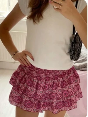 Rosa volangkjol med mönster - Supersöt kort volangkjol i rosa med ett detaljerat mönster. Kjolen har smock i midjan och dubbla volanger. Perfekt för vår och sommar! Kjolen är i xs/xxs💗💗kjolen har inbyggda shorts!