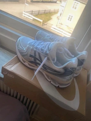 Vita ASICS sneakers med silverdetaljer - Snygga vita ASICS sneakers med silvriga och svarta detaljer. Skorna har en sportig design med mesh- och syntetmaterial för bra andningsförmåga och komfort. Perfekta för dig som gillar en clean och modern stil. Klassisk snörning och låg profil.