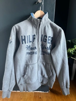 Grå Hilfiger zip hoodie - Snygg grå zip från Tommy Hilfiger med broderad text 'Hilfiger New York Pacific League 1985' på bröstet och ärmen. Zippen har två fickor fram, ribbade muddar och hög krage. Perfekt för en avslappnad look. OBS den är S men passar M