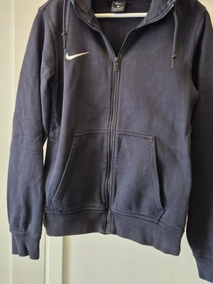 Mörkblå hoodie från Nike - Klassisk mörkblå herr hoodie från Nike med dragkedja, huva med dragsko och två stora fickor framtill. Diskret Nike-logga i vitt på bröstet. Perfekt för en avslappnad och sportig stil.