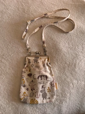 Liten Mumin handväska med axelrem - Söt liten handväska med Mumin-motiv i beige, brunt och gult. Väskan har metallbygel och avtagbar axelrem i tyg. Perfekt för mobilen eller småsaker. Tillverkad i bomullstyg med tryckta figurer från Muminvärlden. Det enda är att axelbandet är lite missfärgat vid noggrann inspektion. Väskan är ca 10x20 cm.