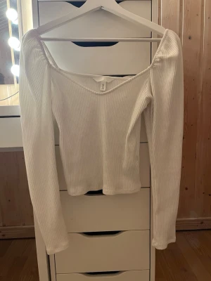 Vit ribbad offshoulder topp - Trendig vit ribbad topp med offshoulder-design och långa ärmar. Snygg v-ringad skärning och stretchigt material som sitter skönt på kroppen. Perfekt att styla med jeans eller kjol för en clean look.