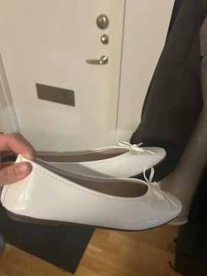 Vita ballerinaskor med rosett - Snygga vita ballerinaskor i glansigt material med rund tå och söt rosettdetalj framtill. Skorna har låg klack och klassisk design som passar till många olika outfits. Perfekta för dig som gillar stilrena och trendiga skor.