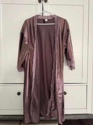 Lila satin morgonrock från Venus - Morgonrock i lila satin från Venus med spetsdetaljer längs kanterna och ärmsluten. Rocken har en mjuk och glansig yta, trekvartsärmar och en elegant, avslappnad passform. Perfekt för att chilla hemma och känna sig lyxig.