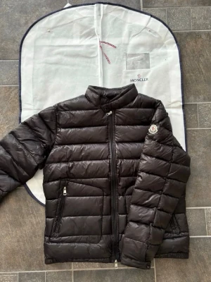 Svart dunjacka från Moncler - Snygg svart dunjacka från Moncler med klassisk quiltad design och ståkrage. Jackan har dragkedja framtill, två sidofickor med dragkedja och Moncler-logga på ärmen. Tillverkad i glansigt material och fylld med dun för extra värme. Perfekt för kalla vinterdagar.