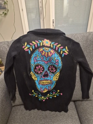 Too Fast lång knitted kofta med  en stor, detaljerad dödskalle i starka färger M - Svart kofta från Too Fast med färgglada broderade motiv på både fram- och baksida. Framsidan har broderade blommor, svalor och hjärtan på fickorna, medan baksidan har ett stort, färgstarkt motiv med blommor. Koftan har långa ärmar och öppen front.