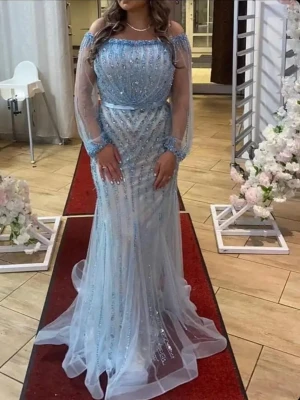 Ljusblå långklänning med paljetter - Elegant ljusblå långklänning med glittriga paljetter och transparenta långa ärmar. Klänningen har offshoulder-modell och markerad midja med ett tunt band. Perfekt för speciella tillfällen och ger en lyxig känsla.