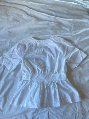 Vit omlottopp med knytband - Snygg vit omlottopp med kort ärm och breda knytband i midjan. Toppen har en djup v-ringning framtill och volangliknande nederdel som ger en feminin siluett. Perfekt att styla med jeans eller kjol för en trendig look.