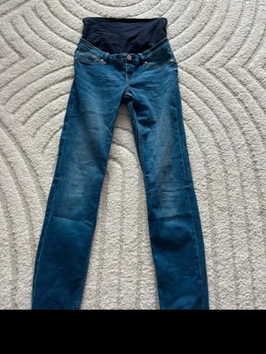 Blå mammjeans med mjuk mudd - Säljer ett par blå mammjeans med bekväm, elastisk mudd över magen. Klassisk femficksmodell med smal passform och snygga slitningar. Perfekta för dig som vill ha både stil och komfort under graviditeten.