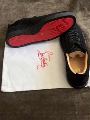 Svarta sneakers från Christian Louboutin - Snygga svarta sneakers från Christian Louboutin med klassisk röd sula och svarta skosnören. Ovandelen är i mocka och insidan är beige med guldfärgad logga. Perfekta för dig som vill ha en exklusiv och stilren look.