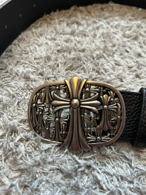Chrome hearts skärp - Snyggt svart bälte med ett stort, detaljerat metallspänne med korsmotiv. Bältet har broderade kors i rött, vitt, gult och rosa längs remmen. Perfekt accessoar för att lyfta din outfit med en unik touch.