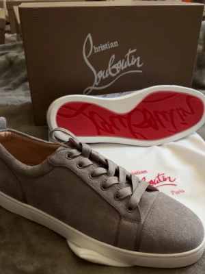 Christian Louboutin gråa sneakers - Snygga gråa sneakers från Christian Louboutin i mocka med klassisk röd sula och diskret logga. Skorna har lågt skaft, rund tå och matchande gråa skosnören. Perfekta för dig som vill ha en lyxig och stilren look.
