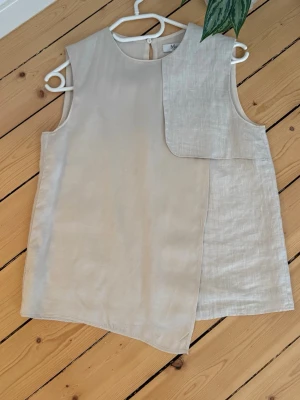 Beige asymmetrisk linnetopp från Marimekko - Snygg beige ärmlös linnetopp från Marimekko med unik asymmetrisk design och lager på lager-detalj framtill. Toppen har rund halsringning och knäppning bak i nacken. Perfekt för dig som gillar minimalistisk och stilren look.
