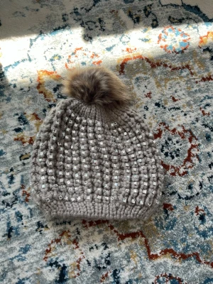 Beige stickad mössa med pälsboll - Snygg beige stickad mössa med glittriga stenar och en fluffig pälsboll på toppen. Insidan är fodrad med mjuk, vit fleece för extra värme. Perfekt accessoar för kalla dagar och en trendig look.