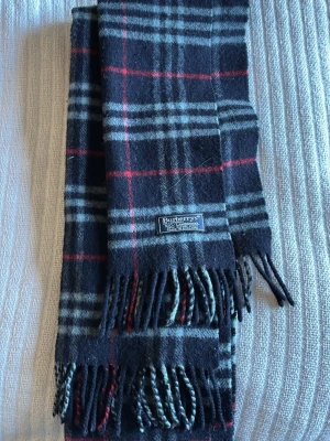 Mörkgrå rutig halsduk från Burberry - Klassisk halsduk från Burberry i mörkgrått med rutigt mönster i vitt och rött. Halsduken har fransar i ändarna och är tillverkad i mjuk ull. Perfekt accessoar för att lyfta din outfit med en tidlös design.