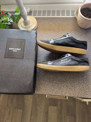 Svarta Saint Laurent sneakers - Svarta sneakers från Saint Laurent med beige sula och handskriven logga på sidan. Skorna har ovandel i canvas och detaljer i läder, klassisk snörning och låg profil. Kommer med originalkartong och dustbag. Skorna har gått upp lite som ni ser på dem 2 sista bilderna men kan fixas lätt hos en sko makare tvätt ingår, mvh Robin🌟