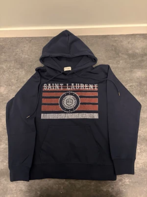 Mörkblå hoodie från Saint Laurent - Cool mörkblå hoodie från Saint Laurent med tryck i vitt och rött på bröstet. stor magficka och ribbade muddar. Materialet är mjukt och bekvämt, perfekt för en avslappnad look. Trycket har en vintagekänsla med SL-logga och stjärnor.