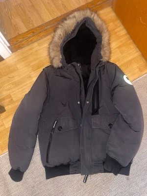 Svart dunjacka med pälskantad huva - Snygg svart dunjacka från Northwest Outfitting  SMOG med pälskantad huva och coolt reflexmärke på ärmen. Jackan har dragkedja, knappar, två stora fickor framtill och ribbade muddar. Perfekt för kalla vinterdagar.