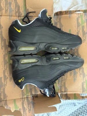 Nike Air Max 95 Svarta/Gula sneakers - Nike Air Max 95 sneakers i svart med gula detaljer och synlig Air Max-dämpning i sulan. Snygg vågig design på ovandelen, broderad gul Swoosh och signaturdetaljer på hälen. Ovandel i mesh och syntetmaterial, klassisk snörning och vadderad krage.