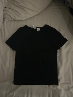 Svart basic t-shirt från Divided - En svart, enkel t-shirt/magtröja från H&M med rund hals och korta ärmar. Perfekt att matcha med jeans eller kjol för en clean och stilren look. Tillverkad i mjukt bomullsmaterial som känns skönt mot huden. 