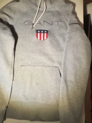 Grå hoodie från GANT med logga - Snygg grå hoodie från GANT med broderad logga och sköld på bröstet. Klassisk känguruficka framtill och vit dragsko i huvan. Mjuk bomullsblandning och sportig känsla, perfekt till jeans eller joggers.