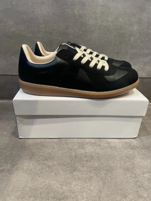 Maison margiela - Snygga svarta sneakers med beige gummisula och vita skosnören. Ovandelen är i en mix av läder och mocka för en stilren look. Insidan är ljus och ger en fräsch kontrast. Perfekta för dig som gillar klassisk och clean stil.