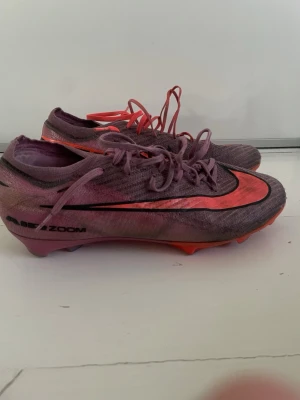 Nike Air Zoom Mercurial rosa fotbollsskor - Säljer ett par Nike Air Zoom Mercurial fotbollsskor i rosa och röd med svarta detaljer. Skorna har låg profil, snörning och en tunn syntetisk ovandel för bra bollkontroll. Sulan har fasta dobbar för spel på gräs. Perfekta för dig som vill ha speed och stil på planen.