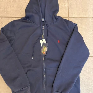 Mörkblå hoodie från Polo Ralph Lauren - Snygg mörkblå hoodie från Polo Ralph Lauren med dragkedja och huva. Klassisk design med röd logobrodyr på bröstet och ribbade muddar. Perfekt för dig som gillar stilrena och bekväma plagg.