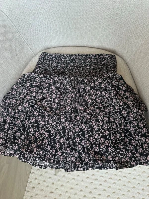 Svart blommig kjol från KappAhl - Supersöt svart kjol från KappAhl med små ljusrosa blommor och smock i midjan. Kjolen har volanger och är gjord i ett lätt, luftigt tyg som ger en skön känsla. Perfekt för dig som gillar en romantisk och trendig stil.