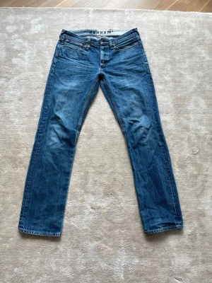 Blå raka jeans från Denham - Säljer ett par klassiska blå jeans från Denham med rak passform och fem fickor. Jeansen har en snygg tvättad look och detaljer som synliga sömmar och läderpatch bak i midjan. Perfekta till en avslappnad stil och tillverkade i slitstarkt denimtyg. (Litet hål i skrevet)