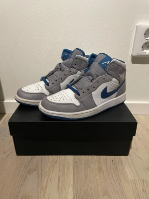 Air Jordan 1 Mid grå/blå sneakers - Snygga Air Jordan 1 Mid sneakers i grått, vitt och blått. Skorna har klassisk siluett med blå swoosh, gråa paneler och vit bas. Tillverkade i läder med Air Jordan-logga på sidan och blå detaljer på plös och sula. Perfekta för dig som gillar streetwear.