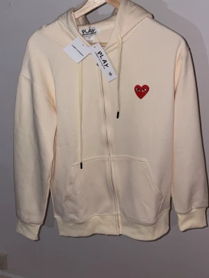 Vit hoodie Comme des Garçons Play - Vit hoodie från Comme des Garçons Play med dragkedja, huva och fickor. Fram finns det klassiska röda hjärtat med ögon och på ryggen flera röda hjärtan samt PLAY- och Comme des Garçons-tryck. Tillverkad i mjukt bomullsmaterial och har snören i huvan.