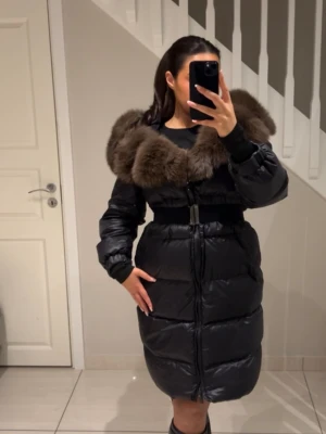 Svart lång dunjacka med pälskrage - Säljer en svart lång dunjacka med stor, fluffig brun pälskrage och markerad midja med bälte. Jackan har dragkedja framtill, quiltad design och långa ärmar. Perfekt för kalla vinterdagar och riktigt snygg siluett.