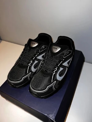 Svarta Dior sneakers med silverdetaljer - Snygga svarta Dior sneakers med mesh och syntetmaterial, silverfärgade detaljer och logga på plösen. Skorna har en chunky sula och coola kontrastsömmar som ger en modern vibe. Perfekta för dig som vill sticka ut med stilrena och exklusiva sneakers.
