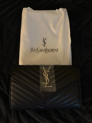 Svart kuvertväska från Saint Laurent - Snygg svart kuvertväska från Saint Laurent i quiltat läder med silverfärgad YSL-logga framtill. Väskan har magnetlås, kedjerem och innerfack med dragkedja. Perfekt för att lyfta vilken outfit som helst.