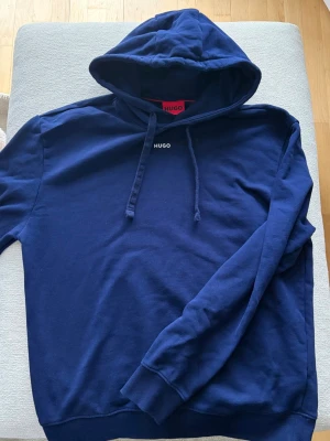 Mörkblå hoodie från Hugo - Snygg mörkblå hoodie från Hugo med minimalistisk logga på bröstet och klassisk huva med dragsko. Tillverkad i mjuk bomullsblandning och har ribbade muddar vid ärmslut och nederkant. Perfekt för en avslappnad och stilren look.
