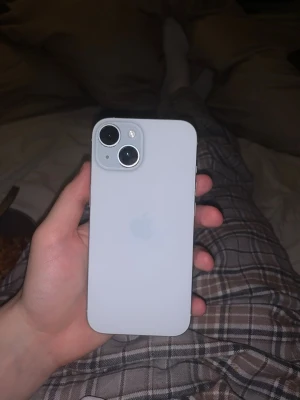 Vit iPhone 15 med dubbla kameror - Säljer en stilren vit iPhone med två stora kameralinser på baksidan och klassisk Apple-logga. Telefonen har rundade hörn och en slät glasbaksida. Perfekt för dig som gillar enkel och modern design.