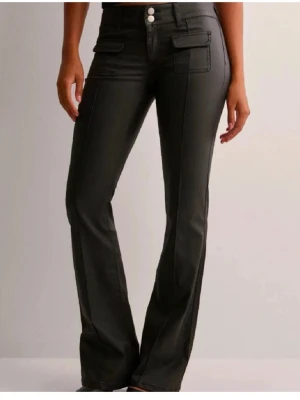 Svarta bootcut byxor Nelly - Snygga svarta skinn byxor med bootcut passform. Byxorna har dekorativa ficklock framtill och dubbla knappar i midjan. Materialet ser ut att vara bomull eller polyester med en lätt glansig finish. Perfekta för en trendig och stilren look.